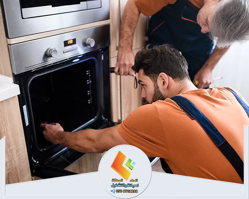 قطع غيار افران الكهرباء بالرياض 0578673748 اتحاد المملكة رجلان يعملان على إصلاح فرن في مطبخ، يظهران التركيز على صيانة أفران جليم غاز بالرياض