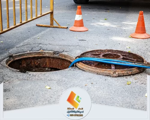 فتحة صرف صحي مفتوحة في الشارع بجوار غطاء معدني وخرطوم مياه أزرق مع حواجز مرورية.